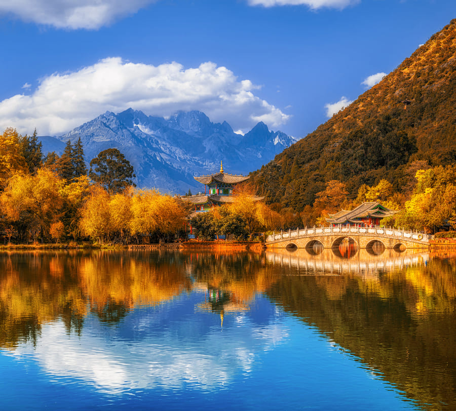 El Estanque del Dragón Negro en Yunnan, China, mostrando un pabellón tradicional y un puente de piedra arqueado rodeados de árboles otoñales dorados, perfectamente reflejados en un lago cristalino con una imponente montaña nevada de fondo bajo un cielo azul.