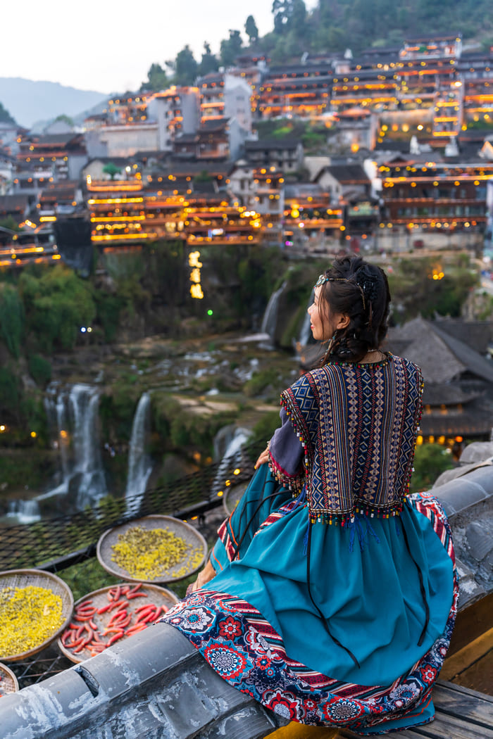 Vista en perspectiva de una mujer de espaldas con un vestido turquesa y chaleco tradicional, contemplando un pueblo iluminado en una colina junto a una cascada.