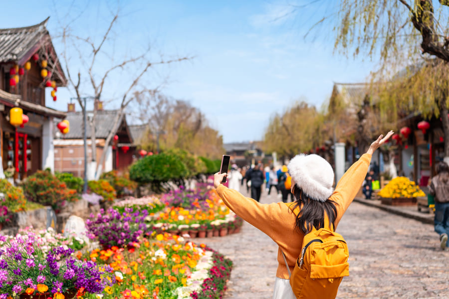Una turista de espaldas con mochila amarilla y gorro blanco levanta los brazos alegremente mientras se toma una foto con su celular en una calle peatonal de un pueblo tradicional chino, rodeada de coloridos parterres de flores y arquitectura antigua.