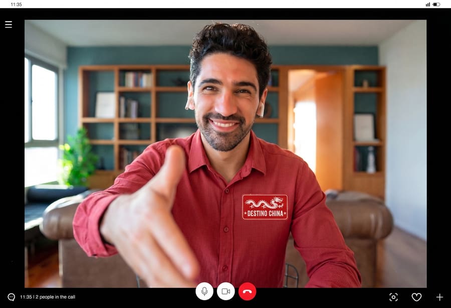 Un hombre sonriente con una camisa roja extiende su mano para estrecharla en una videollamada. Lleva un parche que dice "DESTINO CHINA". Se encuentra en un entorno de oficina en casa acogedor y moderno